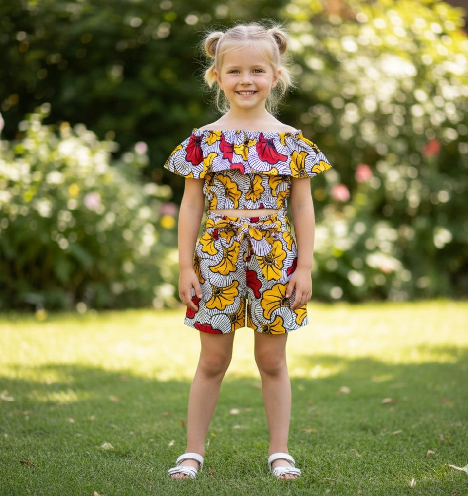 Fillette portant un ensemble en wax fleuri composé d’un crop top à épaules dénudées et d’un short taille haute avec ceinture nouée. Tenue colorée et élégante inspirée de la mode africaine.
