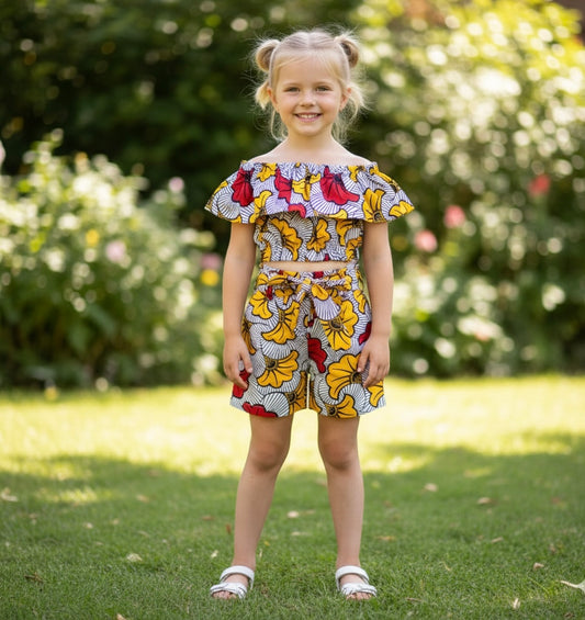 Fillette portant un ensemble en wax fleuri composé d’un crop top à épaules dénudées et d’un short taille haute avec ceinture nouée. Tenue colorée et élégante inspirée de la mode africaine.