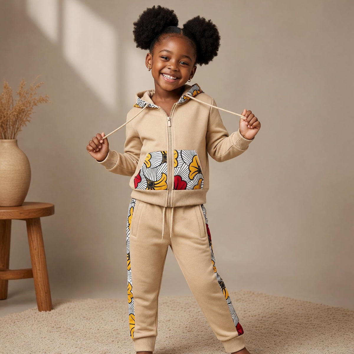 Enfant portant un ensemble jogging MPESA beige avec veste zippée à capuche et détails en wax jaune, rouge, noir et blanc sur les poches et le pantalon.